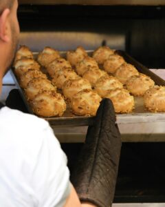 A baker with oven gloves taking a tray of freshly baked bread out of an oven in a bakery.